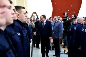 Wiceminister Spraw Wewnętrznych i Administracji gratuluje ściskając dłoń nowoprzyjętym policjantom. Za nim Komendant Wojewódzki Policji w Białymstoku, a w tle rodziny i bliscy nowoprzyjętych policjantów.