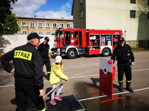 Zdjęcie w kolorze. Na pierwszym planie strażacy oraz dziecko biorące udział w dyscyplinie sportowej.