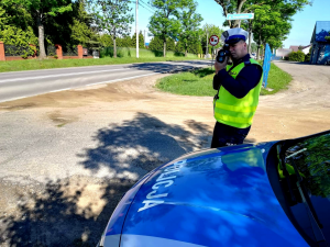 Policjant dokonuje pomiaru prędkości stojąc na poboczu. Obok jest zaparkowany radiowóz Policji.