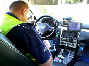 Policjant sporządza dokumentacje służbową siedząc wewnątrz nieoznakowanego radiowozu za kierownicą.