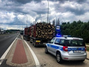 radiowóz policji, a przed nim pojazd ciężarowy z naczepą, na której znajduje się źle zabezpieczone drzewo