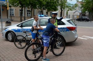 na pierwszym planie 2 chłopców na rowerze, z nimi  policjant ruchu drogowego a za nim policyjny radiowóz