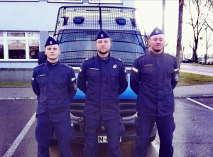 Trzech policjantów z oddziału prewencji policji stoi przed oznakowanym radiowozem marki Mercedes.