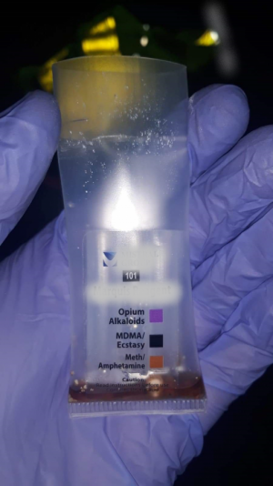 dłoń w niebieskiej rękawiczce trzymająca woreczek plastykowy na woreczku oznaczenia nazw narkotyków przy kolorowych kwadratach w środku brązowa ciecz