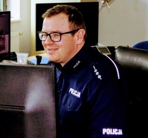 Policjant w trakcie służby. Siedzi przy biurku. Przed nim monitor komputera.