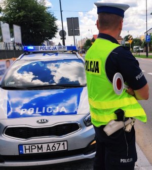 policjant w trakcie służby; stoi przy radiowozie z tarczą do zatrzymywania pojazdów