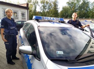 Policjantka i policjant stoją obok radiowozu. Zdjęcie zrobiono w ciągu dnia.