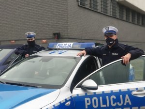 Dwoje policjantów stoi przy radiowozie. Mają maseczki na twarzach.