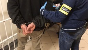 Policjant trzyma zatrzymanego mężczyznę, który ma założone kajdanki na ręce trzymane z tyłu.
