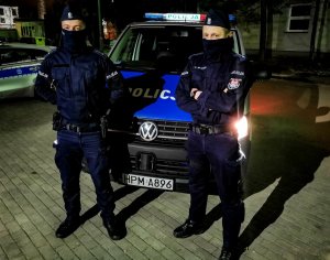 Dwóch policjantów stoi przed radiowozem w porze nocnej.