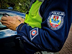 Ręka policjanta, który trzyma w dłoni urządzenie. Na przedramieniu naszywka z logo Komendy Miejskiej Policji w Białymstoku.