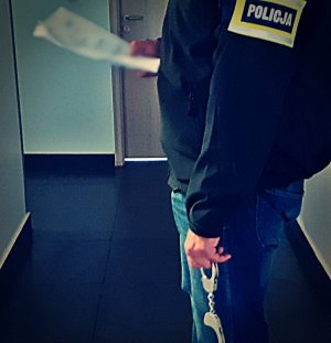mężczyzna z opaska na ramieniu z napisem policja i kajdankami w dłoni stoi na korytarzu
