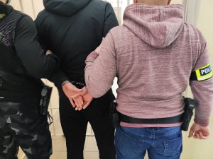 dwaj mężczyźni na ramionach opaski z napisem Policja po środku mężczyzna w czarnej kurtce i kajdankami na nadgarstkach