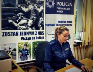 policjantka siedząca przy biurku, za nią bannery o tematyce policyjnej