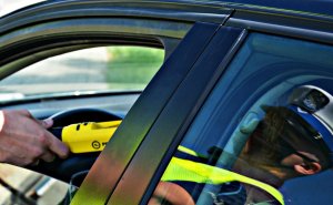 widok na otwarte okno samochodu w oknie dłoń z urządzeniem do badania trzeźwości w tylnej szybie odbicie mężczyzny w mundurze w czapce z białym pokrowcem