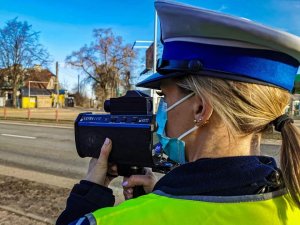 policjantka dokonuje pomiaru prędkości; w tle droga, niebo oraz budynek
