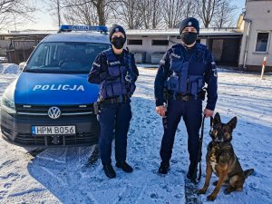 mężczyzna i kobieta w mundurach z napisem policja obok pies w tle samochód z napisem policja na masce, na ziemi śnieg