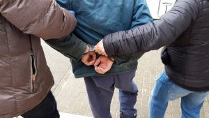 policjanci prowadzą zatrzymanego ; zbliżenie na ręce zatrzymanego w  kajdankach