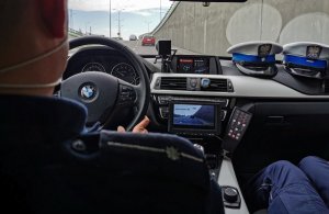 dwóch policjantów wewnątrz radiowozu; przez szybę widać drogie i samochody