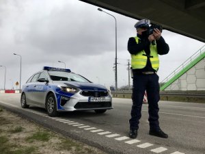 policjant ruchu drogowego dokonuje pomiaru prędkości; na zdjęciu widać też radiowóz i drogę