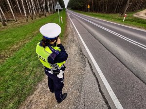policjantka ruchu drogowego dokonuje pomiaru prędkości; w tle: droga, drzewa i pojazdy