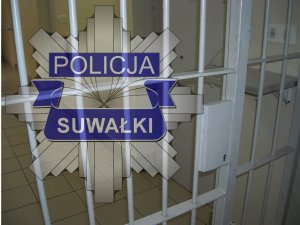 gwiazda policyjna z napisem POLICJA SUWAŁKI; w tle krata od pomieszczenia dla osób zatrzymanych