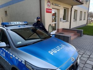 policjant stoi obok radiowozu; na zewnątrz; w tle budynek