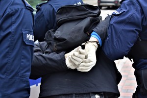 osoba trzymająca ręce za plecami na nadgarstkach kajdanki obok policjanci