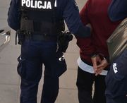 policjant umundurowany trzyma i prowadzi zatrzymanego w kajdankach na ręce trzymane z tyłu