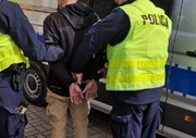 policjanci prowadzą zatrzymanego w kajdankach na ręce z tyłu w tle radiowóz