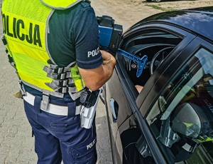 samochód koloru ciemnego z otwartym oknem od strony kierowcy przy nim stoi policjant w mundurze i kamizelce odblaskowej z napisem policja. Trzyma w ręku urządzenie do mierzenia prędkości