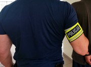 plecy policjanta w granatowej koszulce z opaską na ramieniu policja trzymający za ramę osobę z zielonej bluzie. Wszyscy widziani od tyłu