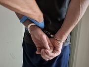 mężczyzna stojący tyłem i mający na nadgarstkach założone kajdanki. Obok widać ręce w gumowych niebieskich rękawiczkach , które trzymają za przedramię rękę w kajdankach.