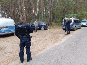 cztery osoby, w tym dwóch policjantów w mundurach stojący na drodze przy lesie. Obok zaparkowane radiowozy policyjne i granatowy samochód osobowy