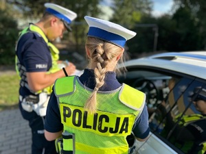 dwóch policjantów ruchu drogowego, kobieta i mężczyzna stojący w mundurach i kamizelkach odblaskowych z napisem policja stojący przy samochodzie osobowym, w którym są otwarte drzwi kierowcy