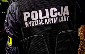 policjant stojący tyłem w kamizelce z napisem białymi literami policja wydział kryminalny  Widać tylko jego plecy i jedną rękę