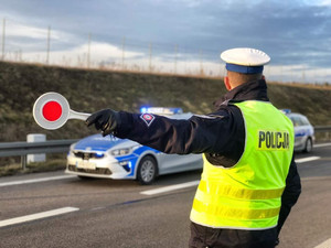 policjant w mundurze widziany od tyłu w kamizelce odblaskowej z napisem policja trzymający w lewej dłoni urządzenie do zatrzymywania samochodów do kontroli a  przed nim stojące dwa radiowozy, jeden za drugim