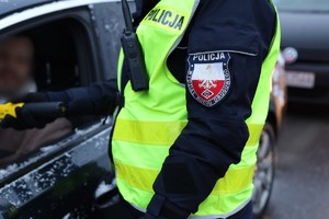 policjant w mundurze i kamizelce odblaskowej z napisem policja stoi przy otwartym oknie samochodu osobowego trzymając w ręku urządzenie do mierzenia zawartości alkoholu w organizmie,które podaje kierowcy siedzącemu w aucie