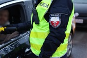 policjant w mundurze i kamizelce odblaskowej z napisem policja stoi przy otwartym oknie samochodu osobowego trzymając w ręku urządzenie do mierzenia zawartości alkoholu w organizmie,które podaje kierowcy siedzącemu w aucie