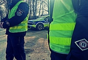 dwóch policjantów w mundurach i kamizelkach odblaskowych z napisem policja a za nimi stojący radiowóz