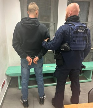 dwie osoby stojące tyłem w pomieszczeniu z kratami w oknach. Jeden mężczyzna to policjant w mundurze, drugi ma zapięte kajdanki na rękach