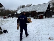 policjant w mundurze widziany od tyłu niesie w rękach drzewo na opał, jest zima i leży śnieg, widać w oddali dach budynku