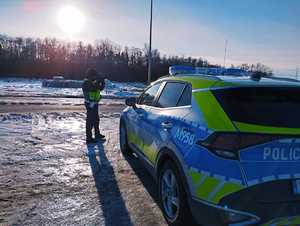 policjant stojący przy jezdni i radiowozie mierzący prędkość urządzeniem do mierzenia