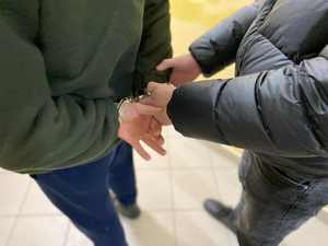 mężczyzna w kajdankach zapiętych na nadgarstkach z tyłu i obok mężczyzna stojący i trzymasjacy za ramię