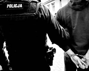 policjant prowadzący za rękę osobę zatrzymaną w kajdankach. Widziani tyłem